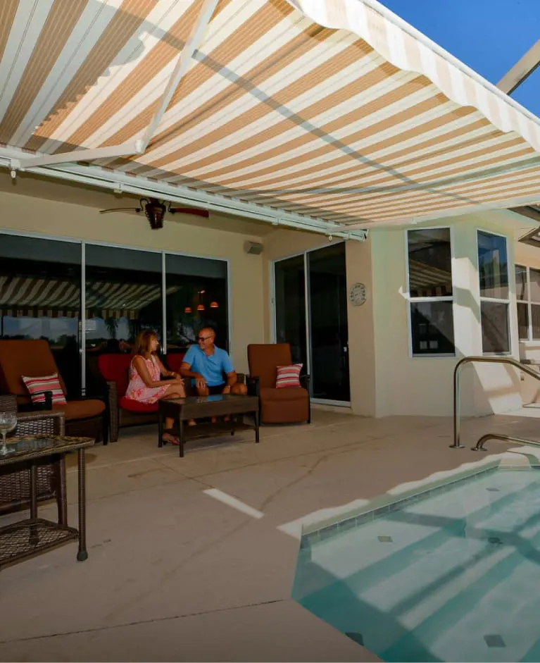 SunPro retractable awning extended over a furnished poolside patio while two people relax in the shade.